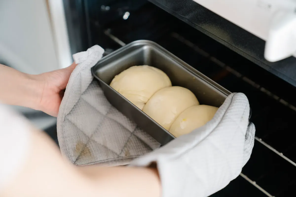 Homemade bread dough placed in oven with glove-covered hands. Fresh baking moment captured.