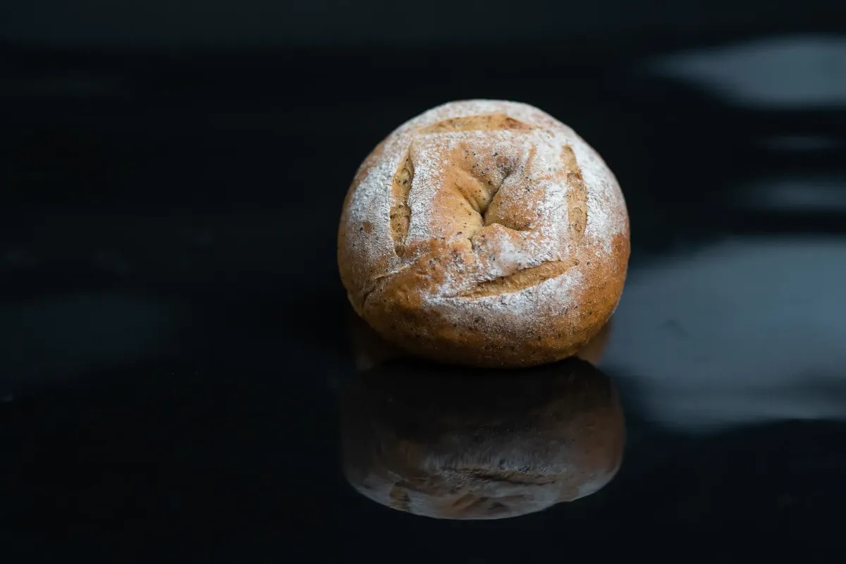 A fresh wheat bun with a crisp golden crust and flour dusting on a reflective black surface.