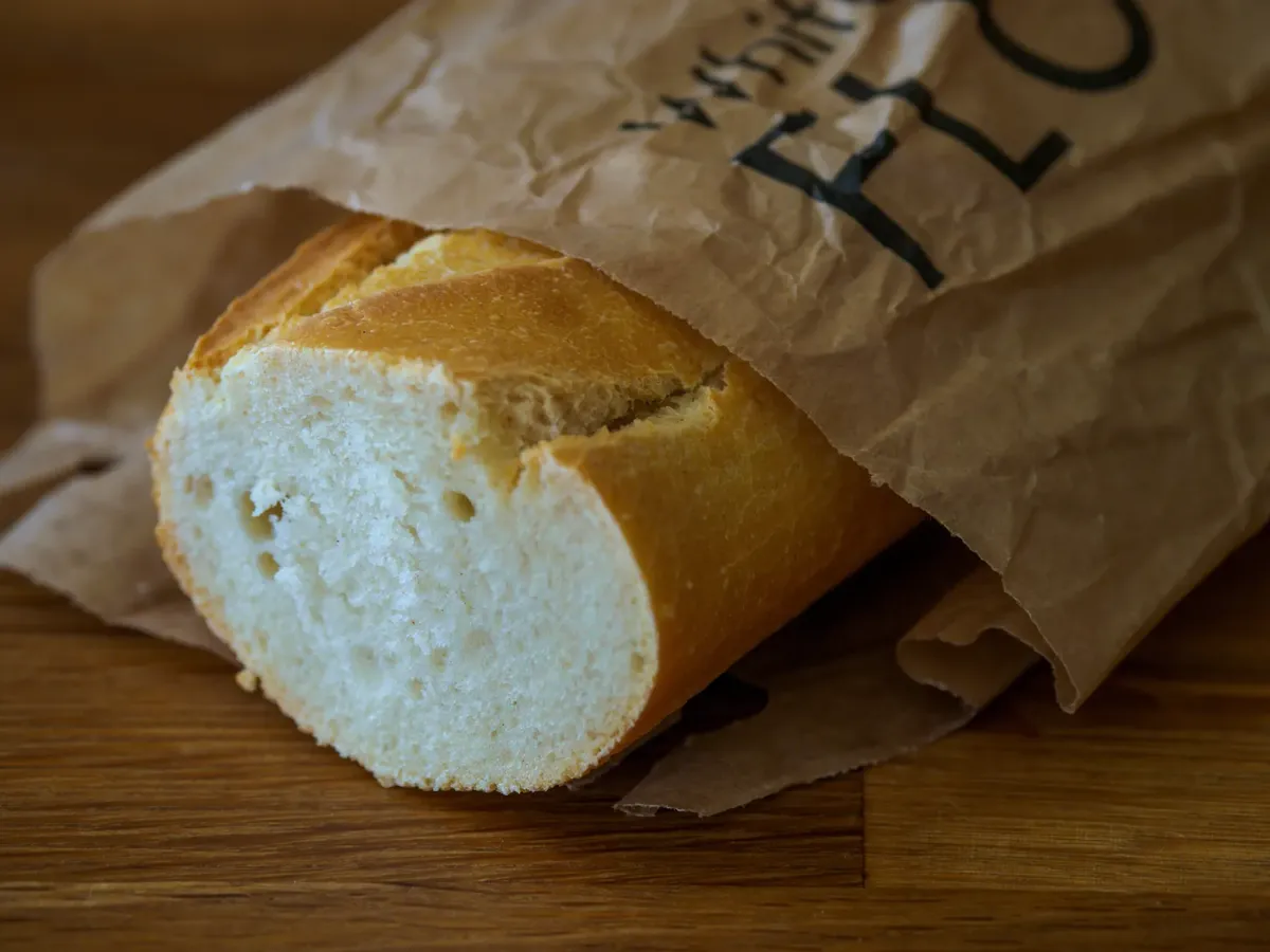 A freshly baked baguette partially wrapped in a paper bag on a wooden surface.