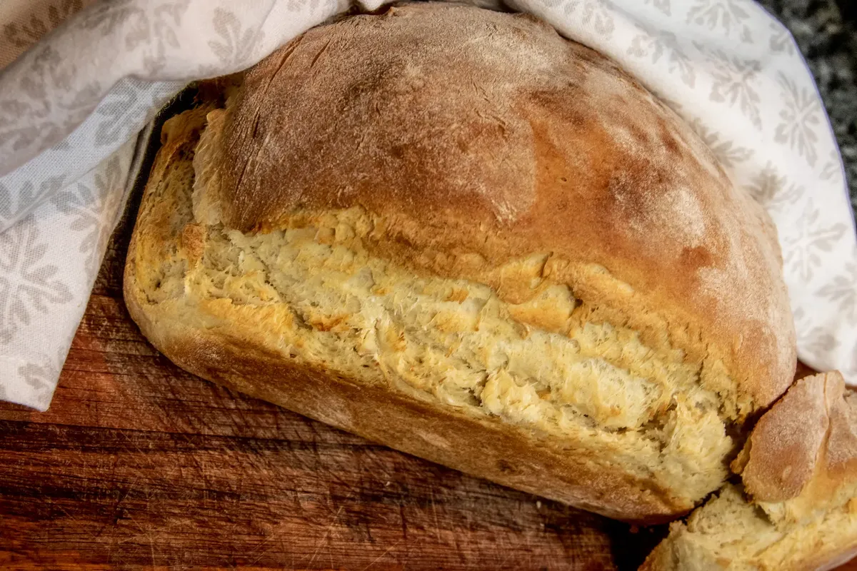 Close-up of a freshly baked homemade bread loaf resting on a wooden board, partially covered by a cloth.