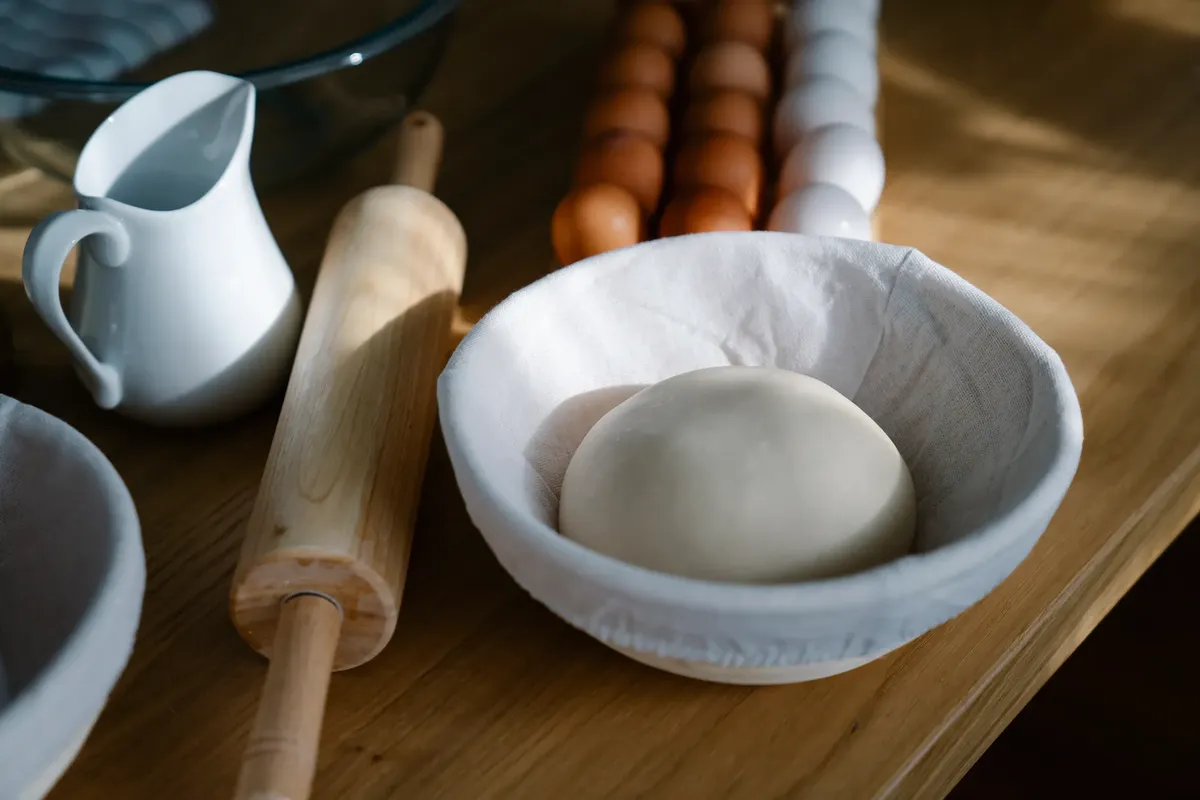 A bowl with dough near a rolling pin, jug, and eggs on a wooden table - perfect for baking themes.